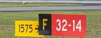 A yellow panel reading “1575 →” butted against a black “F” location panel and a red “32-14” runway designator; the arrow points towards the location message.