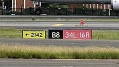 A sign array reading “← 2142” on yellow, “B8” on black, and “34L-16R” on red, standing close behind the runway holding position.