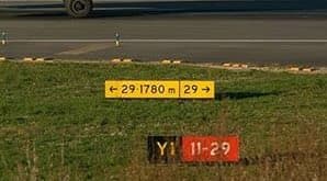 A yellow sign reading “← 29 1780m | 29 →” beside a runway, with a separate yellow “Y1” location panel and red “11-29” designator on the grass in front of it.