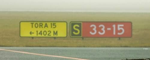 A double-line yellow sign reading “TORA 15” above “← 1402 M”, next to a black “S” location panel and a red “33-15” runway designator.